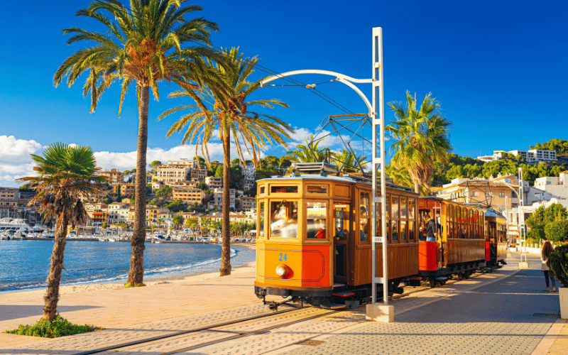 En spårvagn i rörelse som lämnar Port de Sóller, med hamnen, palmträd och husen runt bukten i bakgrunden.